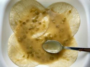 Spoon spreading sauce on layer of corn tortillas in pan for Sour Cream Chicken Enchilada Casserole with zucchini. It's hearty, cheesy, delicious, & easy because everything is layered in one dish.