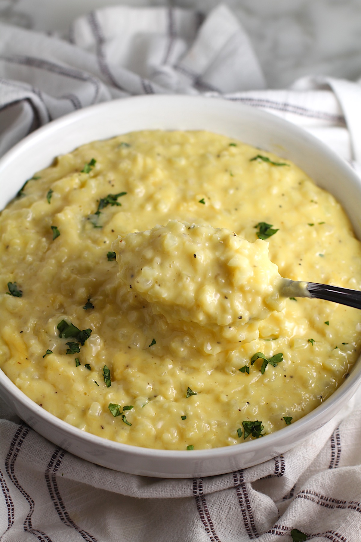 Cheesy Cauliflower Rice with parsley garnish in a bowl with spoon scooping some up. This recipe is so simple with just the flavors from the cauliflower - plus a few other things to help it come together.