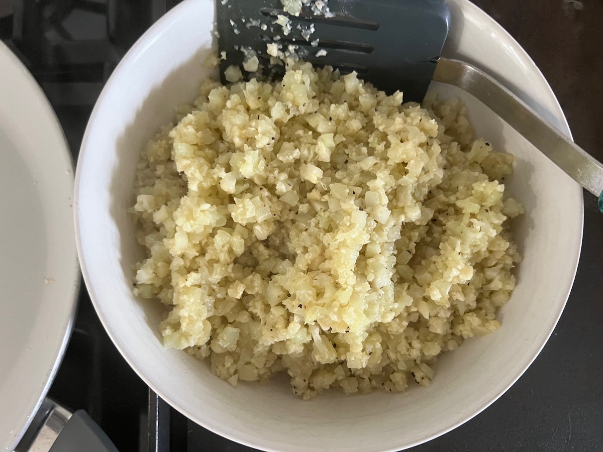 Cooked Cauliflower rice transferred to a bowl next to pan on stove for Cheesy Cauliflower Rice Recipe.