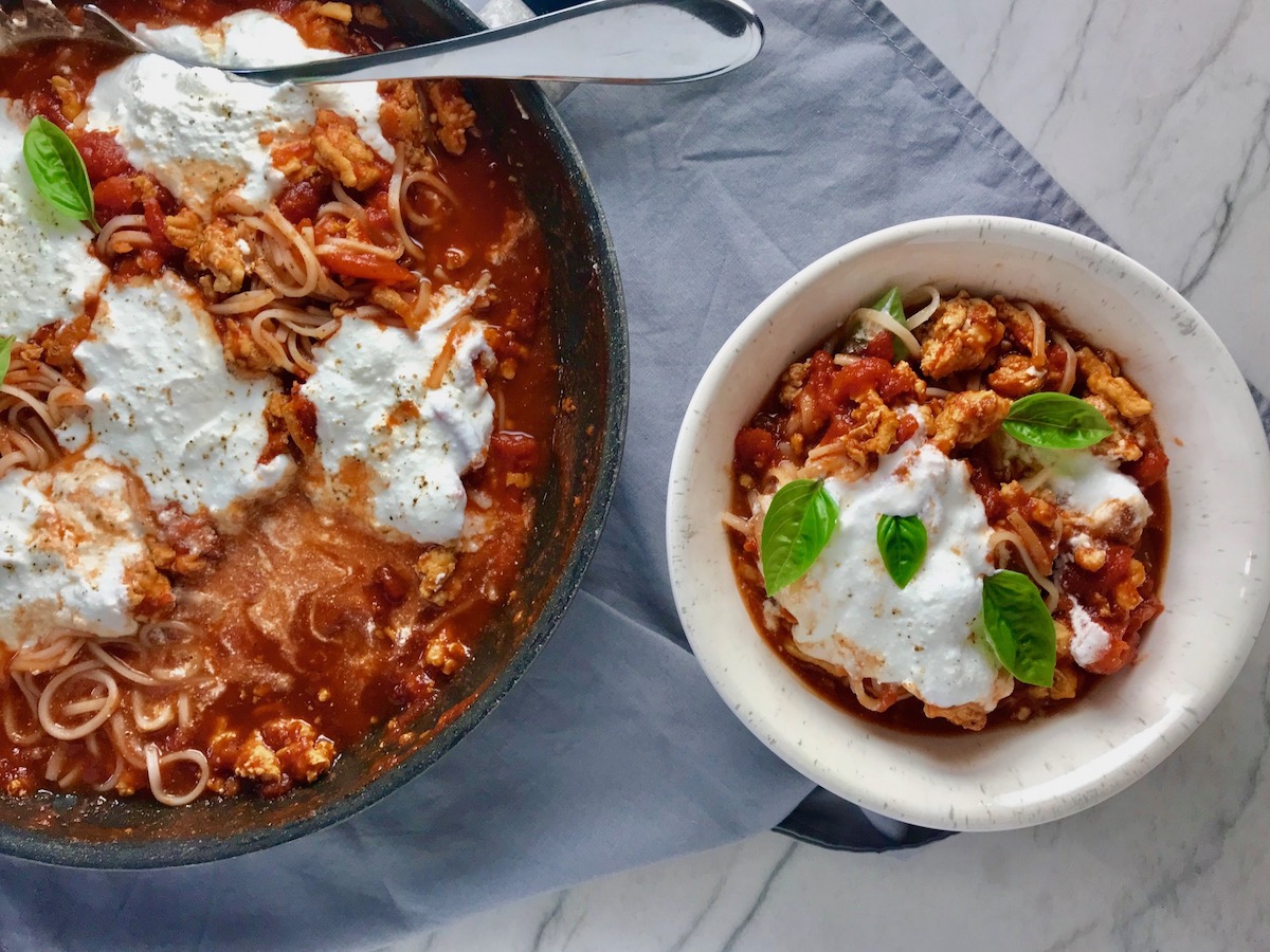 Tomato Chicken Ricotta Pasta in a bowl with fresh basil garnish on counter next to skillet with the rest of the pasta and sauce.