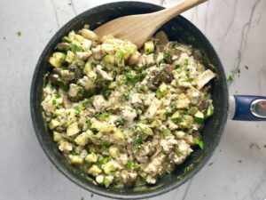 Chicken, Zucchini, Rice Casserole in a skillet on the counter with wood spoon. Skillet has mushrooms, onion, zucchini, chicken, and rice.