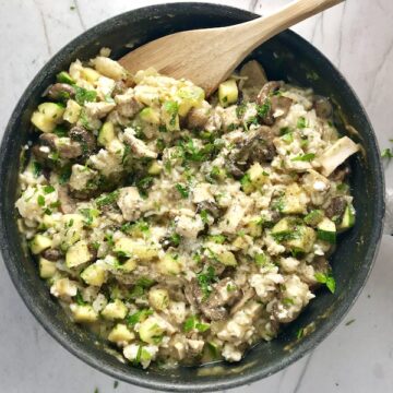 Chicken, Zucchini, Rice Casserole in a skillet on the counter with wood spoon. Skillet has mushrooms, onion, zucchini, chicken, and rice.