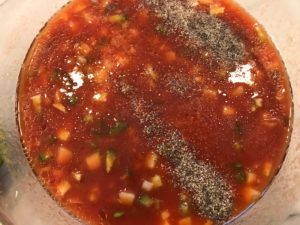 Roasted Red Pepper Gazpacho recipe in a bowl with salt and pepper on top before mixing. The other ingredients are roasted red pepper, green pepper, cucumber, garlic and scallions.