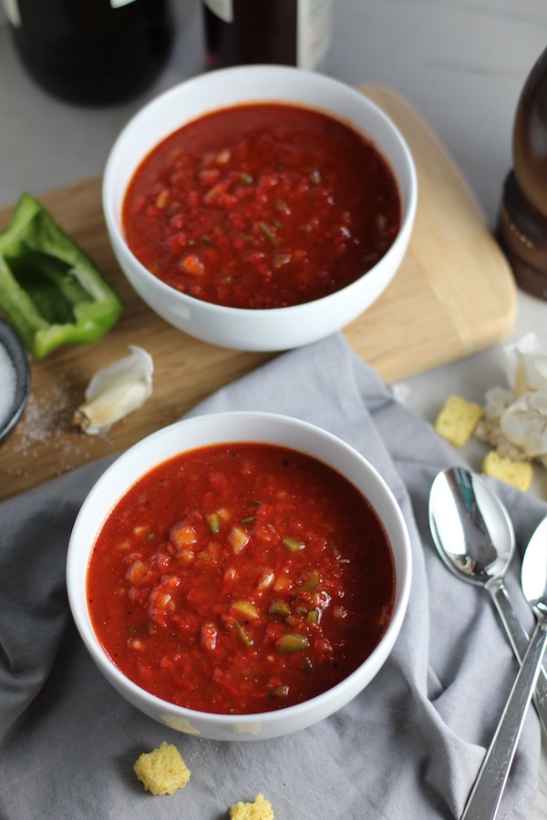 Two white bowls of Roasted Red Pepper Gazpacho recipe from talkingmeals.com. Bowls on napkin and wood cutting board with spoons to the right, green pepper and garlic on cutting board.