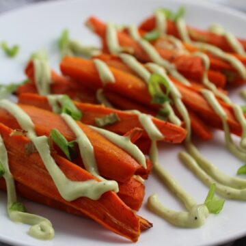 Close up of roasted carrot fries on plate with avocado crema drizzled on top with scallion slices. Roasted Carrots with smoky cumin and scallions, then topped with Avocado Crema are such a delicious and quick side dish for dinner. Roasting brings out the natural sugar in the carrots, so they get a sweet caramelization with the salty and smoky flavors. The Avocado crema gives creamy and silky balance.