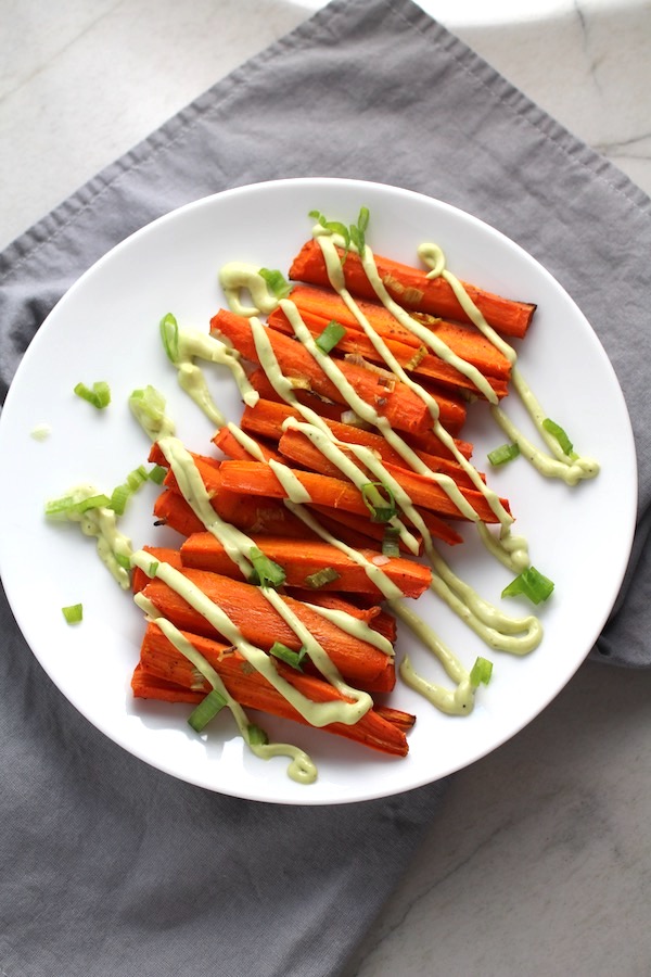 Overhead picture of roasted carrots on plate on gray napkin with avocado crema drizzled on top with scallion slices. Roasted Carrots with smoky cumin and scallions, then topped with Avocado Crema are such a delicious and quick side dish for dinner. Roasting brings out the natural sugar in the carrots, so they get a sweet caramelization with the salty and smoky flavors. The Avocado crema gives creamy and silky balance.