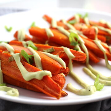 Close up of roasted carrots on plate with avocado crema drizzled on top with scallion slices. Roasted Carrots with smoky cumin and scallions, then topped with Avocado Crema are such a delicious and quick side dish for dinner. Roasting brings out the natural sugar in the carrots, so they get a sweet caramelization with the salty and smoky flavors. The Avocado crema gives creamy and silky balance.