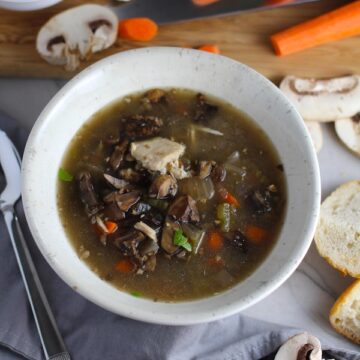 Chicken and Mushroom Soup in white bowl with raw mushrooms, carrots, wood cutting board, bread, and spoon on counter. This Slow Cooker Chicken and Mushroom Soup is warm, hearty, comforting, earthy, and just devine. The mix of blended and chunky mushrooms with carrots, onions, celery, shredded chicken, and herbs gives you a perfect bite every time.