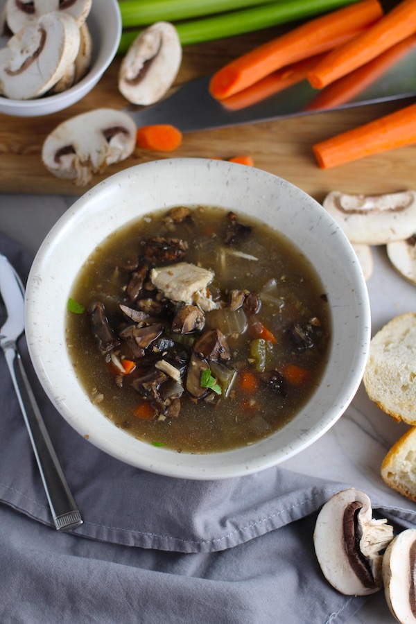 Chicken and Mushroom Soup in white bowl with raw mushrooms, carrots, wood cutting board, bread, and spoon on counter. This Slow Cooker Chicken and Mushroom Soup is warm, hearty, comforting, earthy, and just devine. The mix of blended and chunky mushrooms with carrots, onions, celery, shredded chicken, and herbs gives you a perfect bite every time.