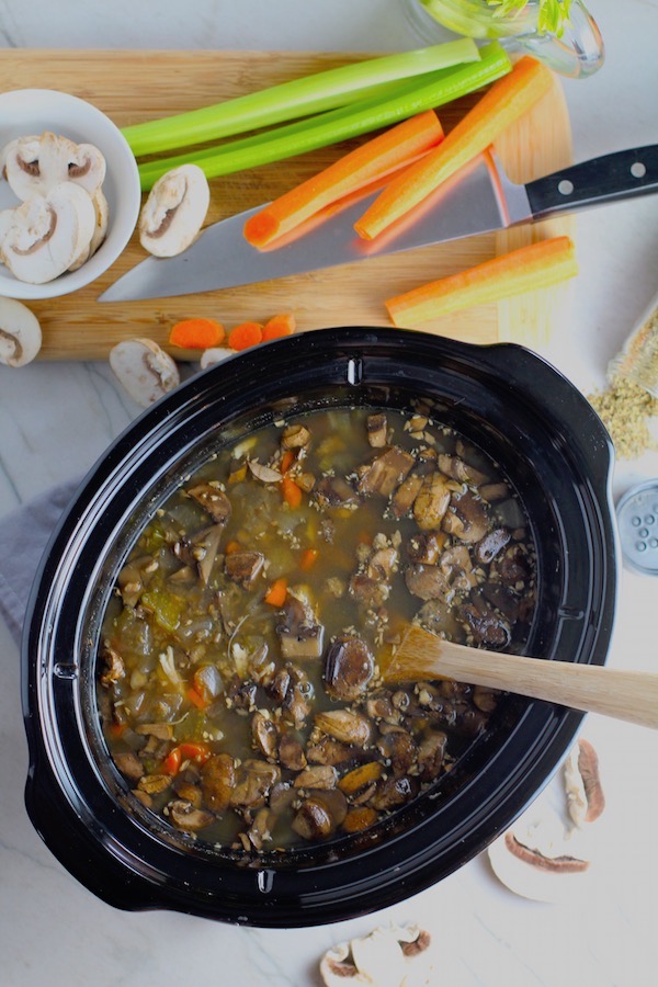 Chicken and Mushroom Soup in a Slow Cooker Pot with wood spoon on counter with cutting board next to it and raw mushrooms and carrots around. This Slow Cooker Chicken and Mushroom Soup is warm, hearty, comforting, earthy, and just devine. The mix of blended and chunky mushrooms with carrots, onions, celery, shredded chicken, and herbs gives you a perfect bite every time.