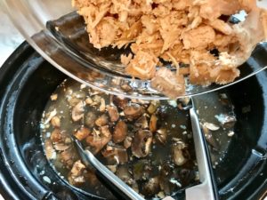 Shredded chicken being added to mushroom soup in a slow cooker with tongs sitting in the soup. This Slow Cooker Chicken and Mushroom Soup is warm, hearty, comforting, earthy, and just devine. The mix of blended and chunky mushrooms with carrots, onions, celery, shredded chicken, and herbs gives you a perfect bite every time.