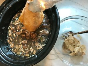 Chicken breast being taken from slow cooker soup with tongs with bowl next to it with another chicken. This Slow Cooker Chicken and Mushroom Soup is warm, hearty, comforting, earthy, and just devine. The mix of blended and chunky mushrooms with carrots, onions, celery, shredded chicken, and herbs gives you a perfect bite every time.