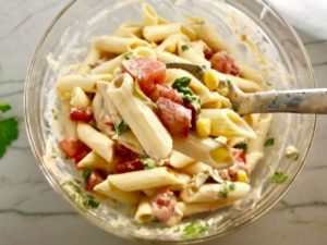 Clear bowl on counter with mixed Mexican Pasta Salad with corn, tomatoes, pasta, Mexican dressing, and cilantro. This Creamy Mexican Pasta Salad has Sun Dried Tomatoes, Corn, Fresh Diced Tomatoes, Cilantro, and smoky Mexican spices. The dressing is creamy and smoky with lots of depth and a hint of sweetness from the Sun Dried Tomatoes that are blended in. Terrific as a side or mix in some chicken or beef and call it a meal!