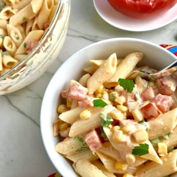 Close up of Mexican Pasta Salad in white bowl on counter with second bowl in upper left corner and tomato half on plate. This Creamy Mexican Pasta Salad has Sun Dried Tomatoes, Corn, Fresh Diced Tomatoes, Cilantro, and smoky Mexican spices. The dressing is creamy and smoky with lots of depth and a hint of sweetness from the Sun Dried Tomatoes that are blended in. Terrific as a side or mix in some chicken or beef and call it a meal!