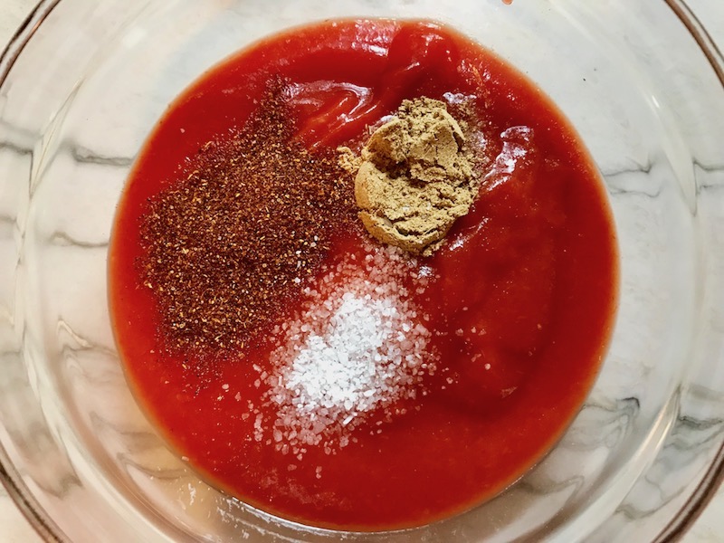Tomato sauce in clear bowl on counter with spices on top: chili powder, cumin, and salt. This Taco Casserole has corn tortillas layered with browned ground chicken seasoned with smokey mexican spices, black beans, tomatoes, and cheese, and then more cheese.