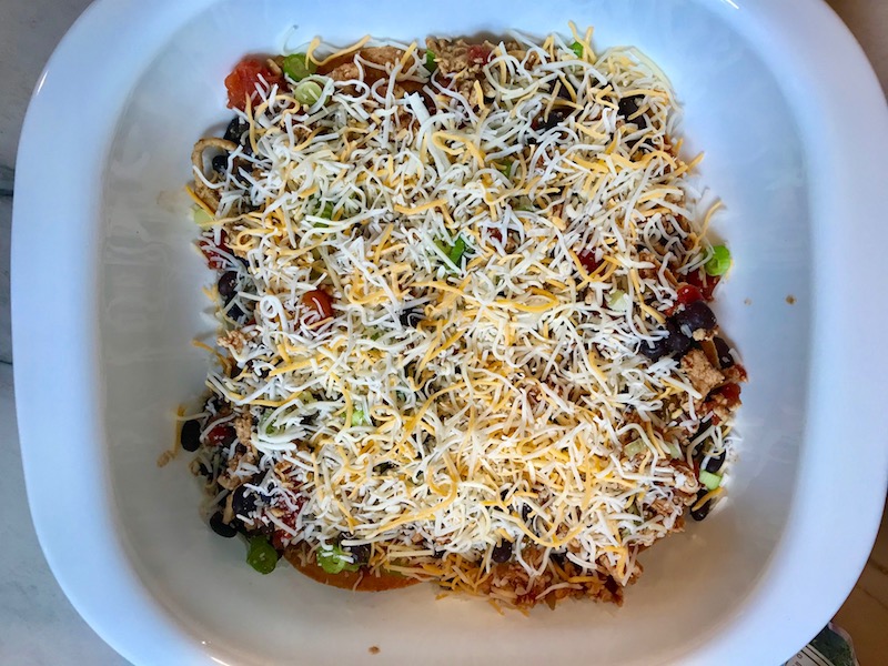 Casserole Dish with the start of the Chicken Taco Casserole Recipe being assembled. Tortillas and ground meat, black beans, tomatoes, and scallions on top..