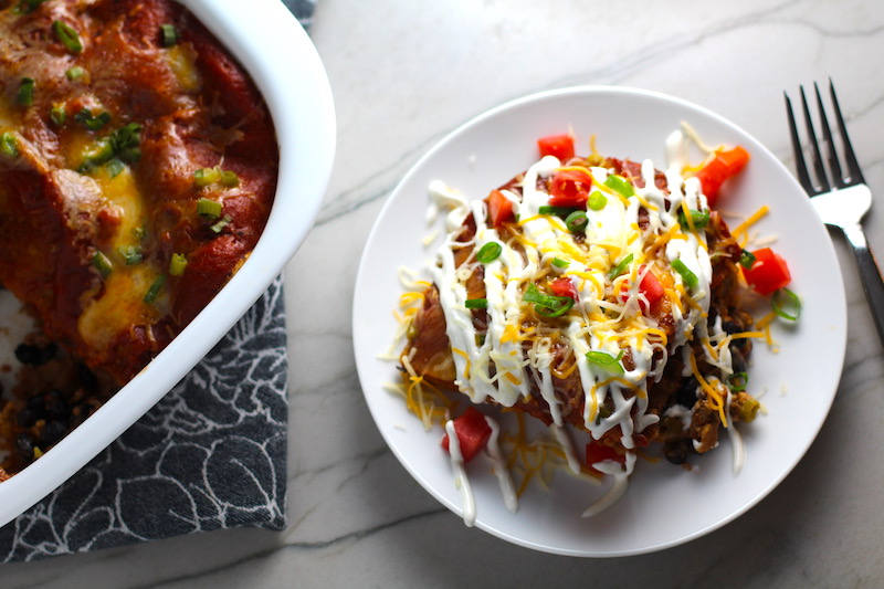 Piece of Taco Casserole on a white plate with sour cream drizzled on top with diced tomato and scallion. Casserole dish to the left on counter. This Taco Casserole has corn tortillas layered with browned ground chicken seasoned with smokey mexican spices, black beans, tomatoes, and cheese, and then more cheese.