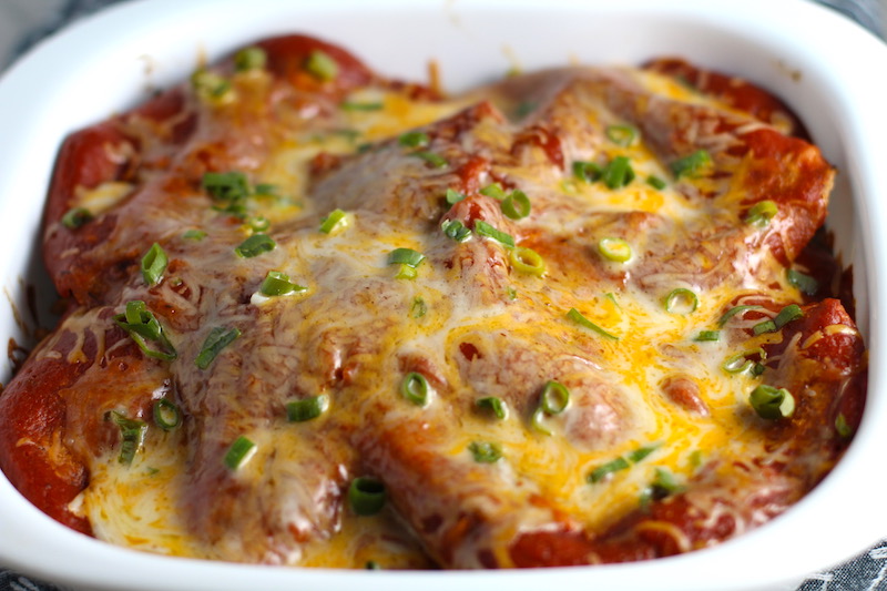 Extreme close up picture of top of taco casserole with red sauce topped with melted cheese and scallion slices. What could be better than tacos? A Taco Casserole that's made with healthier lean ground chicken and can be made ahead for your busy schedule. This Taco Casserole has corn tortillas layered with browned ground chicken seasoned with smokey mexican spices, black beans, tomatoes, and cheese, and then more cheese.