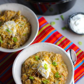 2 Bowls of Chipotle Chicken Enchilada Stew with sour cream and scallion slices as garnish and slow cooker pot in background. It's one of my Prepped Freezer Meal recipes and it's creamy, cozy, hearty, cheesy, and slightly spicy. All ingredients freeze raw, thaw, then cook in the slow cooker or in the oven. It's a stew because it's thick and hearty with bites of chicken, carrots, onion, chipotle peppers, cumin, garlic and tortilla chips that melt down to thicken this delicious stew.