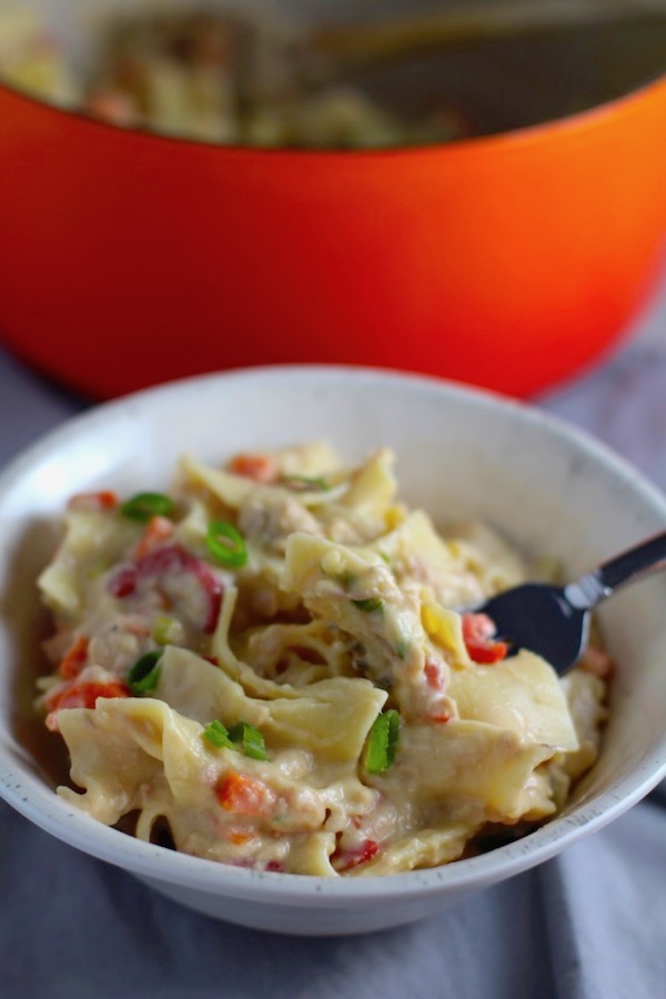 Cheesy Tuna Noodle Casserole Recipe in a bowl with a fork with pot in background. This is a creamy and delicious comfort dish with tuna and noodles and a surprise pop of sweet from Pimiento. This easy recipe is completely from scratch (no canned soup here!) but I can promise you it's so easy and only takes 30 minutes because it's a Casserole that you can make entirely on the stove top!