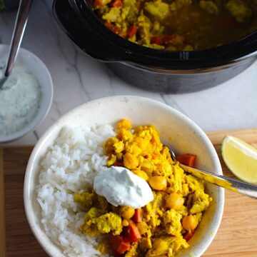 Moroccan Chicken Chili in bowl over white rice with a dollop of Cilantro Lime Yogurt on top and in a bowl to the left. The slow cooker is in background with chili. Moroccan Chicken Chili with Chickpeas is loaded with comforting and healthy ingredients and spices. You get warmth from Ginger, Turmeric, Garlic, and Coriander. Then tons of hearty flavor from browned ground chicken and Shallots. And creaminess from the Carrots and Chickpeas. You can make it ahead and freeze it until you are ready to cook it!