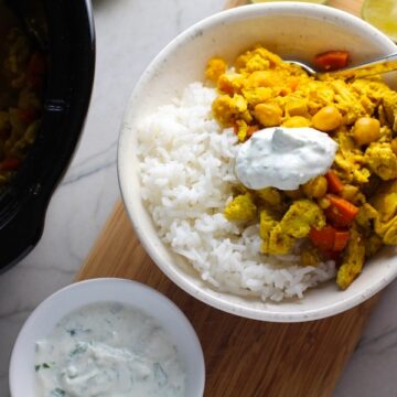 Moroccan Chicken Chili in bowl over white rice with a dollop of Cilantro Lime Yogurt on top and in a bowl to the left. The slow cooker is in background with chili. Moroccan Chicken Chili with Chickpeas is loaded with comforting and healthy ingredients and spices. You get warmth from Ginger, Turmeric, Garlic, and Coriander. Then tons of hearty flavor from browned ground chicken and Shallots. And creaminess from the Carrots and Chickpeas. You can make it ahead and freeze it until you are ready to cook it!