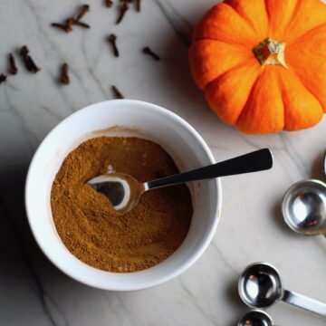 Pumpkin Spice blend in white bowl with silver spoon on counter with mini pumpkins, cloves, and measuring spoons. This Pumpkin Spice Recipe has 4 simple ingredients and takes minutes to make! PLUS, here are 20 EASY WAYS TO USE this Spice blend, many with no baking at all!