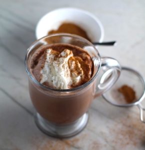 Pumpkin Spice sprinkled on top of whipped cream on hot cocoa. Spice blend in a sifter to the right and in a bowl in background. Recipe has 4 simple ingredients and takes minutes to make! PLUS, here are 20 EASY WAYS TO USE this Spice blend, many with no baking at all!