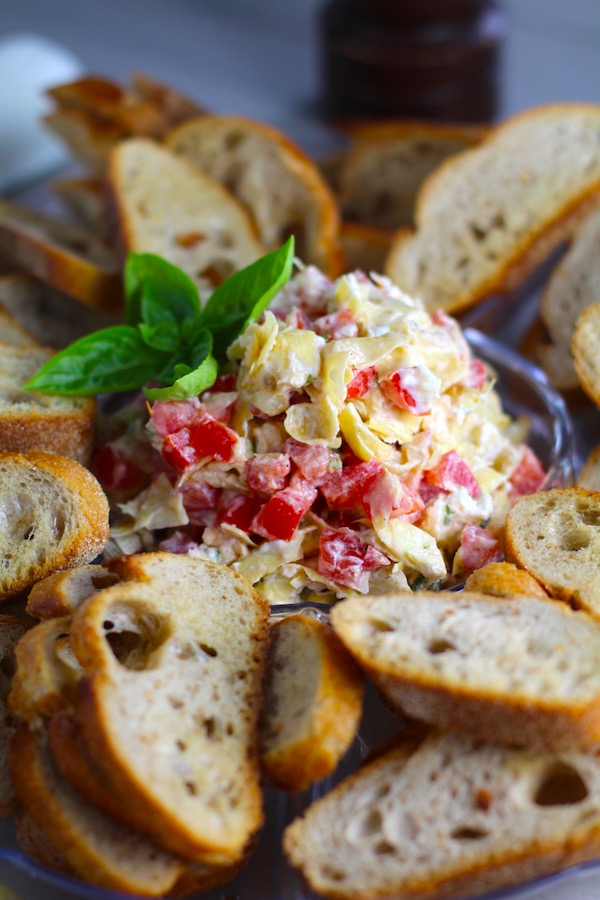 Bruschetta Topping surrounded by crostini on a platter with one crostini topped on a plate. It combines fresh tomatoes, artichoke hearts, garlic, basil, olive oil, and GOAT CHEESE! Top on toasty garlic crostini and you get fresh bright flavors with both crunch and creamy in every bite.