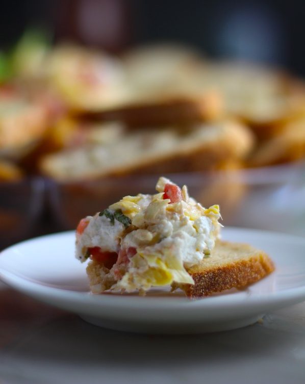 Close up of Bruschetta topping on crostini with platter in background.It combines fresh tomatoes, artichoke hearts, garlic, basil, olive oil, and GOAT CHEESE! Top on toasty garlic crostini and you get fresh bright flavors with both crunch and creamy in every bite.