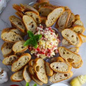 Bruschetta Topping surrounded by crostini on a platter with one crostini topped on a plate. It combines fresh tomatoes, artichoke hearts, garlic, basil, olive oil, and GOAT CHEESE! Top on toasty garlic crostini and you get fresh bright flavors with both crunch and creamy in every bite.