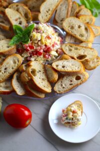 Bruschetta Topping surrounded by crostini on a platter with one crostini topped on a plate. It combines fresh tomatoes, artichoke hearts, garlic, basil, olive oil, and GOAT CHEESE! Top on toasty garlic crostini and you get fresh bright flavors with both crunch and creamy in every bite.