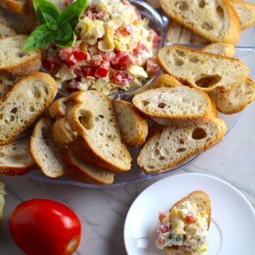 Bruschetta Topping surrounded by crostini on a platter with one crostini topped on a plate. It combines fresh tomatoes, artichoke hearts, garlic, basil, olive oil, and GOAT CHEESE!  Top on toasty garlic crostini and you get fresh bright flavors with both crunch and creamy in every bite.