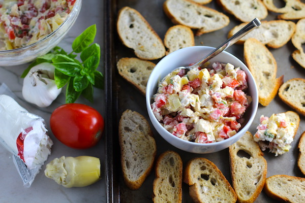 Bruschetta Topping in bowl on pan surrounded by crostini with ingredients to the left. It combines fresh tomatoes, artichoke hearts, garlic, basil, olive oil, and GOAT CHEESE! Top on toasty garlic crostini and you get fresh bright flavors with both crunch and creamy in every bite.