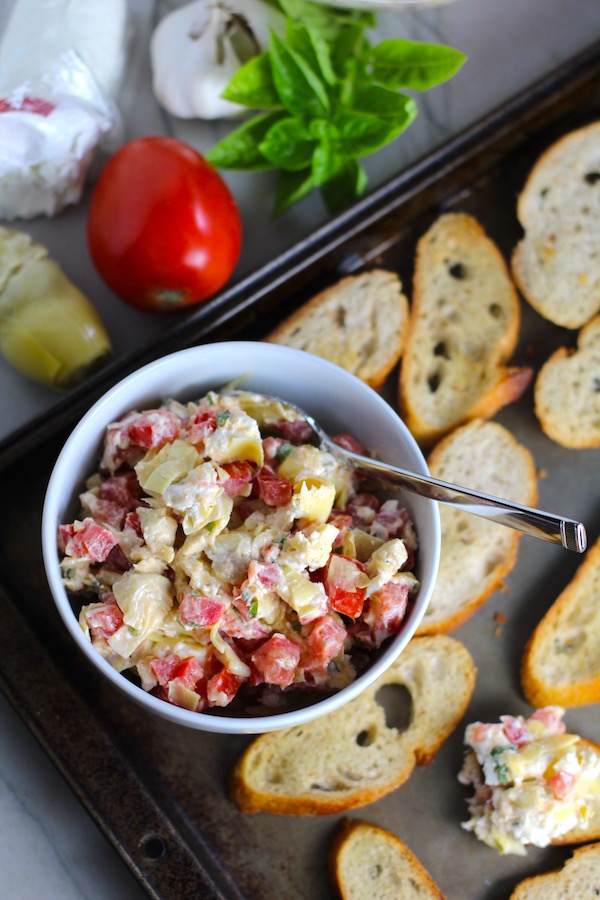 Bruschetta Topping in bowl on pan surrounded by crostini with ingredients to the left. It combines fresh tomatoes, artichoke hearts, garlic, basil, olive oil, and GOAT CHEESE! Top on toasty garlic crostini and you get fresh bright flavors with both crunch and creamy in every bite.