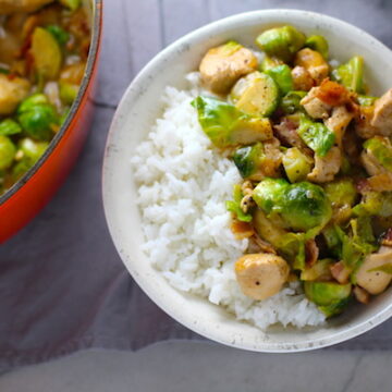 Easy Bacon Brussel Sprouts and Chicken served over white rice in a bowl with orange pot in background. It is a perfect quick Fall Recipe! It has a creamy sauce filled with salty bacon, earthy and almost nutty seared brussel sprouts, and hearty healthy chicken. Serve over rice (or Quinoa or pasta!).