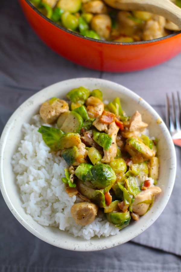 Chicken Brussel Sprouts Bacon served over white rice in a bowl with orange pot in background. It is a perfect quick Fall Recipe! It has a creamy sauce filled with salty bacon, earthy and almost nutty seared brussel sprouts, and hearty healthy chicken. Serve over rice (or Quinoa or pasta!).