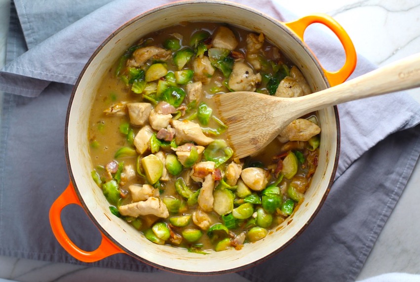 Chicken, Brussels Sprouts, and Bacon in a pot with wood spoon on counter. It's a perfect quick Fall Recipe! It has a creamy sauce filled with salty bacon, earthy and almost nutty seared brussel sprouts, and hearty healthy chicken. Serve over rice (or Quinoa or pasta!).