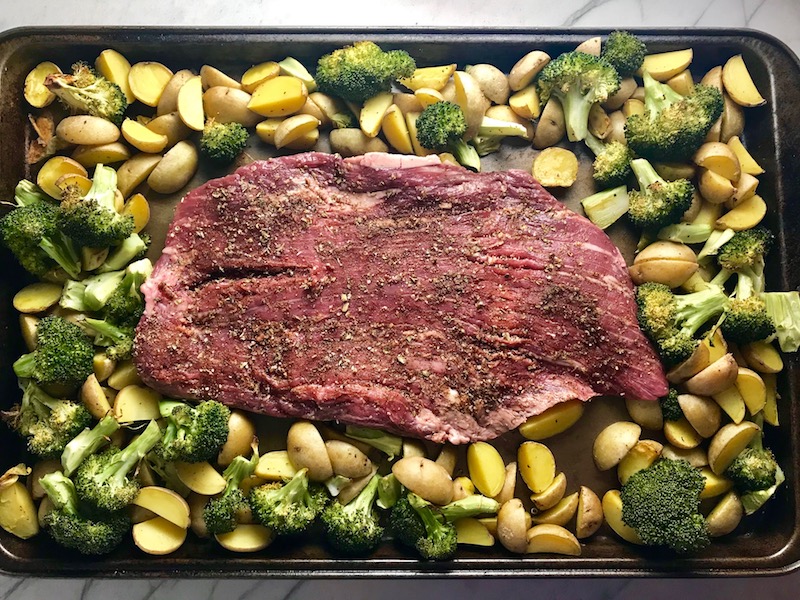 Raw flank steak on sheet pan in center of potatoes and broccoli. This Flank Steak Recipe with Potatoes & Broccoli is an easy and delicious Sheet Pan Dinner. It has a flavorful rub and a tangy, light Garlic Chive Crema!