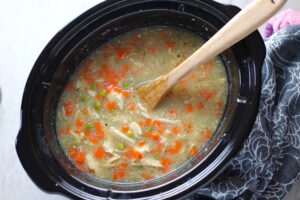 5-Ingredient Homemade Chicken Soup in a crockpot with wood spoon. This soup is thick, creamy, and hearty. It's filled with flavorful broth, meaty shredded chicken, and sweet bursts of bright veggies. You can prepare all of the ingredients ahead and freeze them until you are ready to cook. Then, thaw, pour into the Slow Cooker, set and forget.