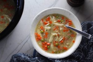 5-Ingredient Homemade Chicken Soup in a bowl with spoon and crockpot in background. This soup is thick, creamy, and hearty. It's filled with flavorful broth, meaty shredded chicken, and sweet bursts of bright veggies. You can prepare all of the ingredients ahead and freeze them until you are ready to cook. Then, thaw, pour into the Slow Cooker, set and forget.