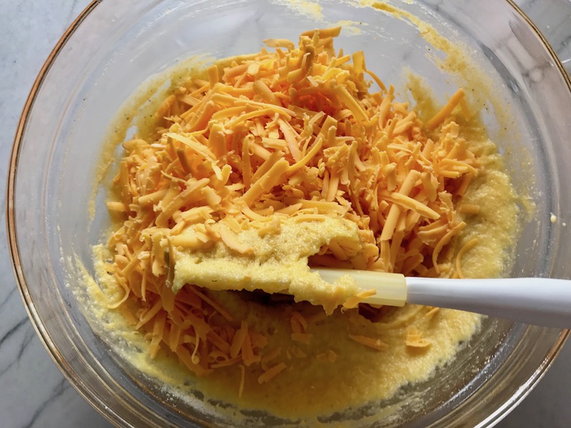 Folding cheese into ingredients in bowl for Cheddar Honey Cornbread piece in hand with more in background. It brings the salty and sweet combo to life spectacularly - particularly when the aromas from the honey, corn, and cheddar fill your house. This Gluten Free version is moist, slight salty from the cheese, slightly sweet from the honey and cornmeal. It's the perfect holiday meal side, whether it's breakfast, brunch, or dinner.