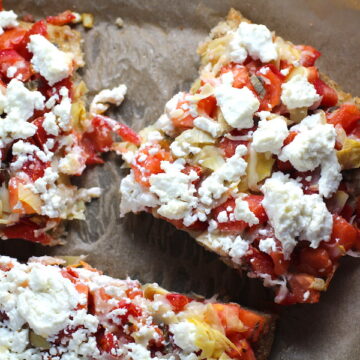 Close up of cut Tomato Tart on parchment. It gives you crunch from the Quinoa crust, a burst of fresh from the tomatoes, tons of flavor from the basil and garlic, tang and meatiness from the artichoke and so much creaminess from the mozzarella and goat cheese! It's addictive and delicious!