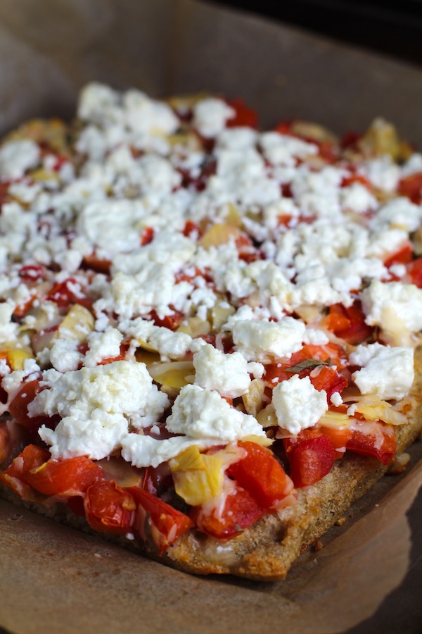 Close up of Tomato Tart corner on parchment. It gives you crunch from the Quinoa crust, a burst of fresh from the tomatoes, tons of flavor from the basil and garlic, tang and meatiness from the artichoke and so much creaminess from the mozzarella and goat cheese! It's addictive and delicious!