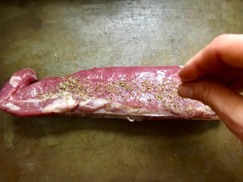 Fingers sprinkling seasoning on raw pork tenderloin on pan for the Caramelized Onions & Mushrooms on Pork Tenderloin recipe. This is such a flavorful main dish! The pork tenderloin is moist and juicy and topped with Onions & Mushrooms that are sauteed low and slow with a touch of balsamic to bring out all of the natural sugars and caramelize them to perfection.