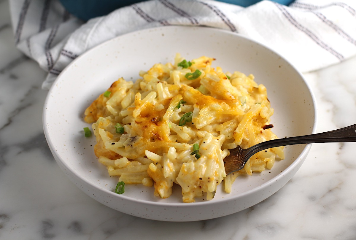 Golden brown cheesy Hash Brown Casserole without Soup scooped out onto a plate with fork on counter with casserole dish in the background.