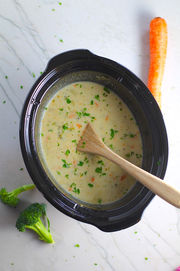 Slow Cooker Creamy Broccoli Soup in a crockpot with wood spoon and raw broccoli and carrots on counter. This is easy, comforting, and delicious! With Carrots, Broccoli, Onion, Garlic, and Oregano, this is one flavorful soup. And, there is no heavy cream, just milk and a little half and half mixed with cornstarch, so it's healthy and gluten free!