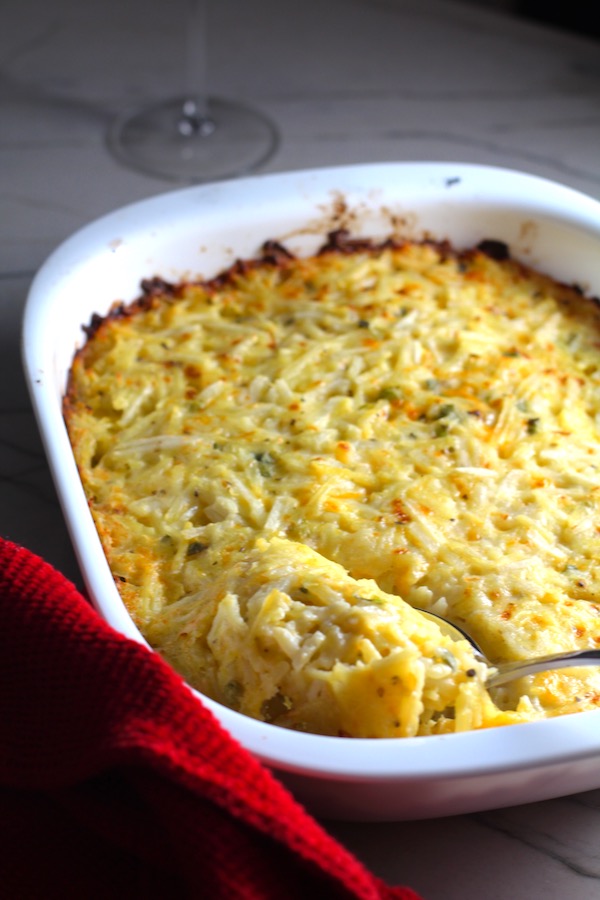 Spoon scooping Cheesy Hash Brown Casserole in casserole dish on counter with red towel. This recipe does not disappoint! It's warm, creamy, and full of flavor! And it's the perfect side dish for your holiday dinner.