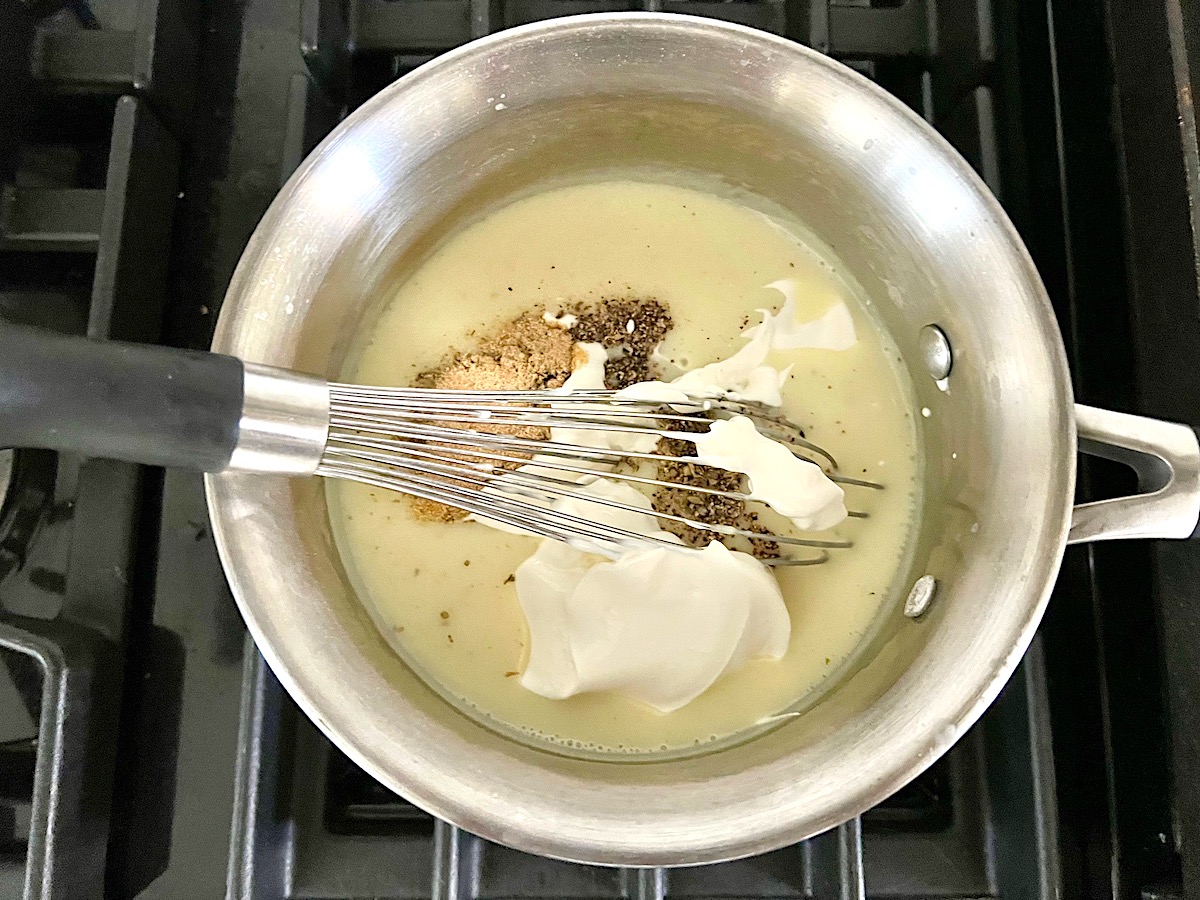 Sour Cream and seasonings being whisked into the sauce in a saucepan on the stove for Hash Brown Casserole without Soup.