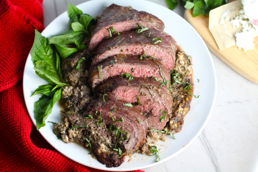 Stuffed Flank Steak cooked and cut on white plate with stuffing oozing out and basil on side. In the stuffing you get mushrooms, onions, and garlic sauteed together. Then they are mixed with fresh parsley and basil, then parmesan, and goat cheese! Roll it all up in Flank Steak and roast for an amazing dinner.